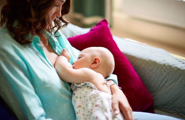 Een moeder met een blauw shirt aan geeft haar baby borstvoeding in een zittende houding op een comfortabele, grijze bank.