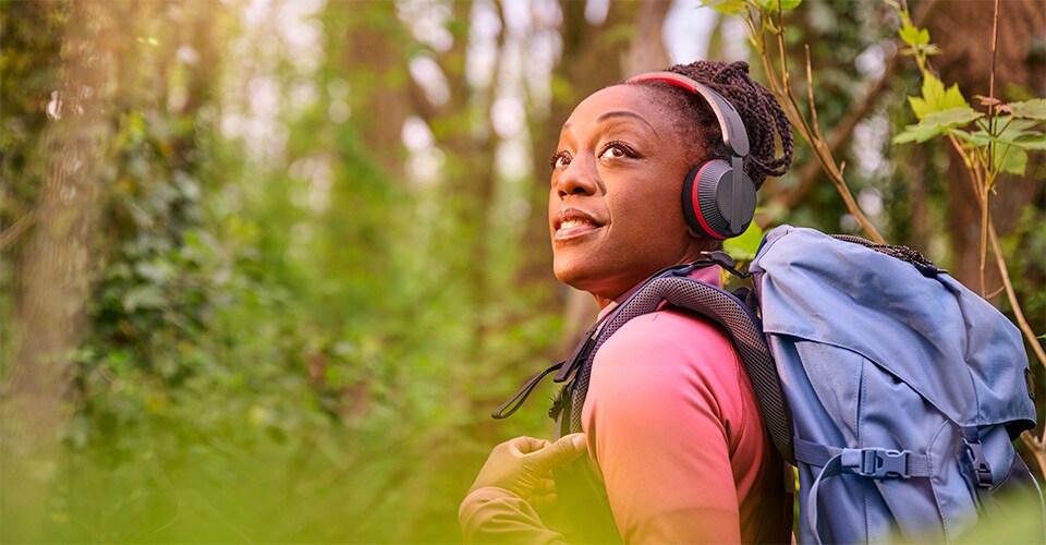Une femme fait de la randonnée dans une forêt avec un sac à dos bleu, portant un casque Philips A6219 et levant les yeux avec un sourire