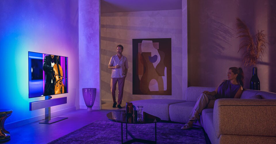 Man en vrouw in een stijlvolle woonkamer kijken naar een Philips Ambilight-tv waarop een cello-optreden wordt getoond