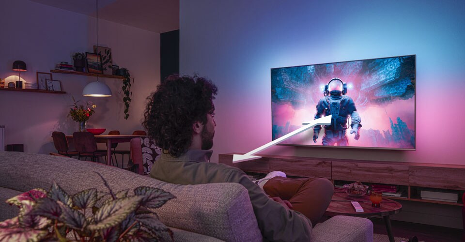 Man zit op een bank en kijkt naar een Philips Ambilight-tv met een kleurrijke astronautscène, waarbij een pijl de optimale kijkafstand aangeeft.