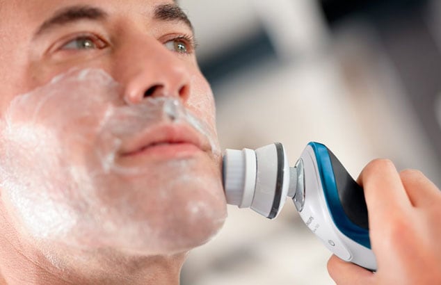 Close-up van een man met scheerschuim op zijn gezicht die een wit, blauw en grijs elektrisch scheerapparaat gebruikt.