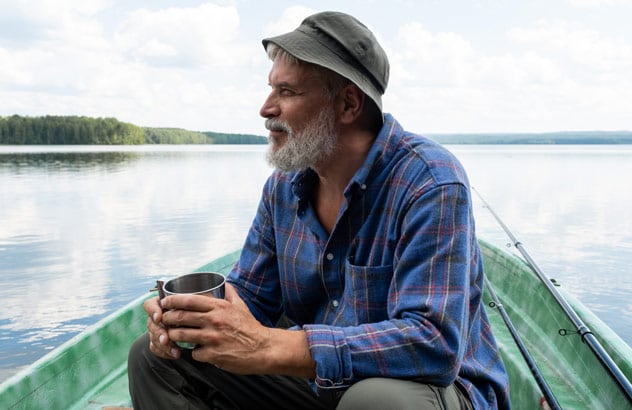 Een oudere man met een grijze baard, een vissershoed en een blauw geruit overhemd zit op een boot en kijkt in de verte.