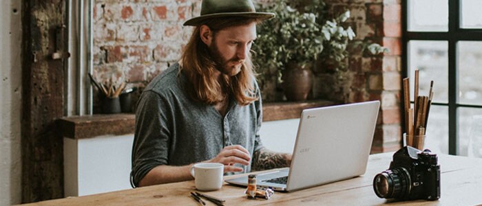 Jonge man met lang haar en een volle baard zittend in een hippe ruimte kijkend naar een laptop voor hem.