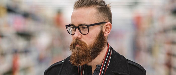 Man met kort haar en goed getrimde baard met zwarte bril voor een wazige winkelachtergrond.