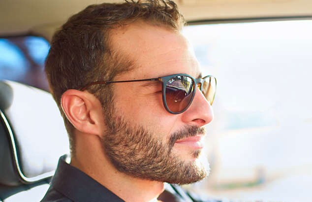 Man met een zwarte zonnebril, een volle bruine baard met snor en een kort kapsel bestuurt een auto.