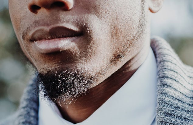 Close up van een zwarte man in een wit overhemd en grijs vest en zijn kinbaard.