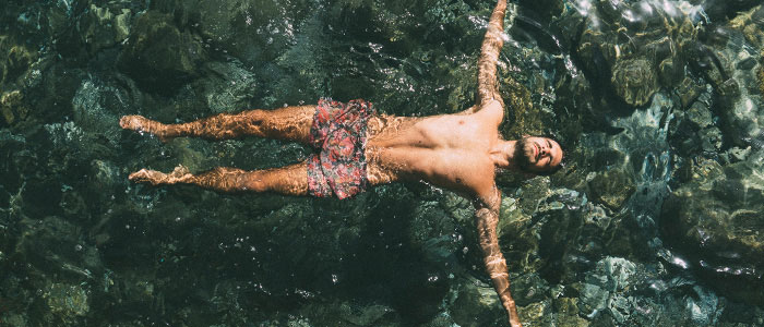 Breed shot van een bebaarde man in zwemshort die met zijn gezicht omhoog in het water drijft met zijn armen gespreid.