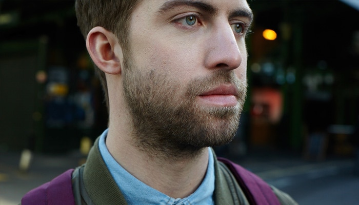 Bruinharige man met een baard van 1 cm die gezicht en nek bedekt, staat in een straat en draagt een paarse rugzak.